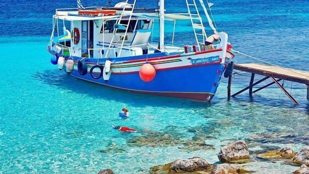A boat docked at a clear beach with rocky foreground and mountains in the background. - Medusa Studios (Kefalonia)