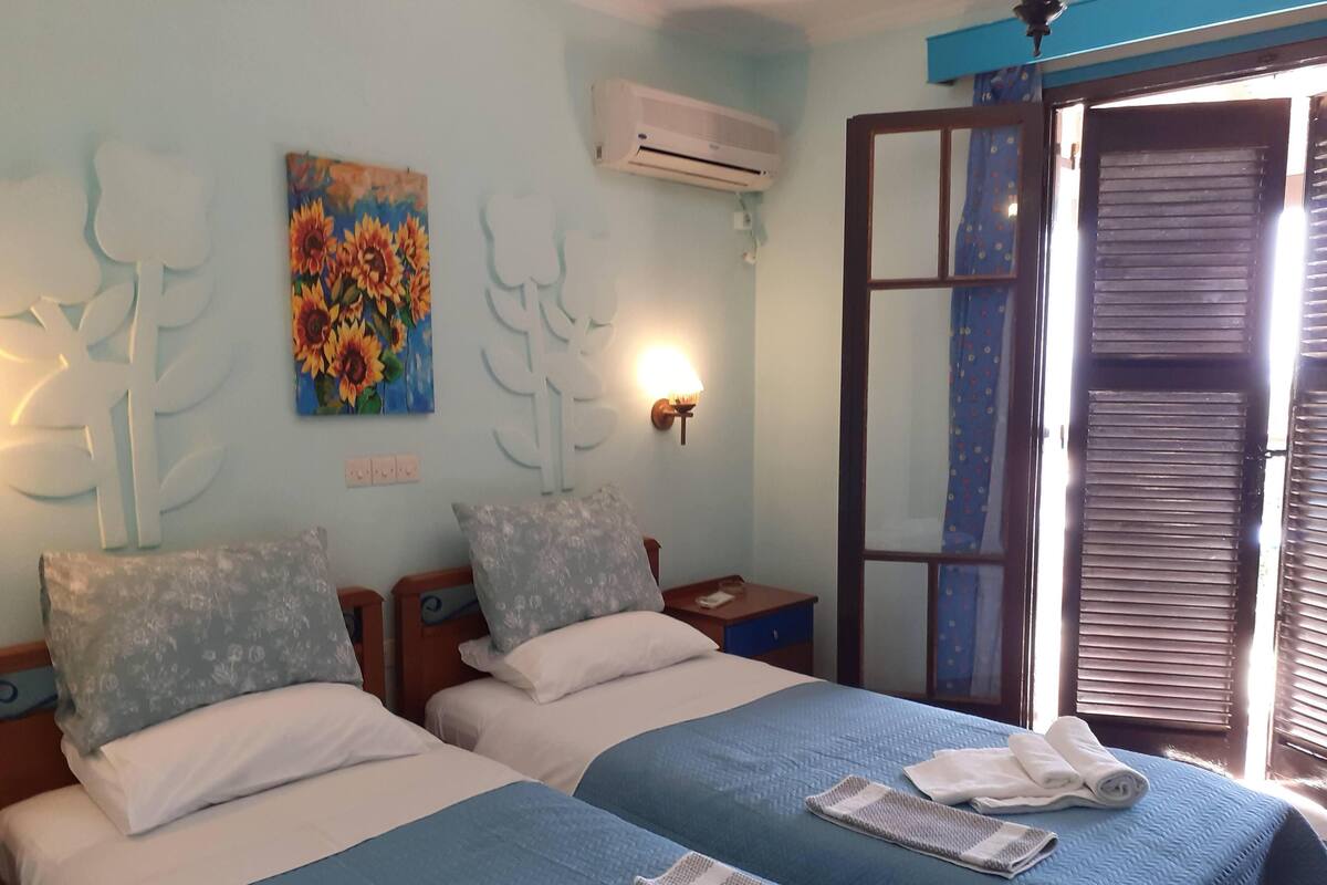 A hotel room with two beds, a painting of sunflowers, an air conditioning unit, and a window with blinds.