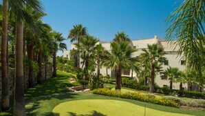 A golf course with a putting green, surrounded by palm trees and a building in the background.