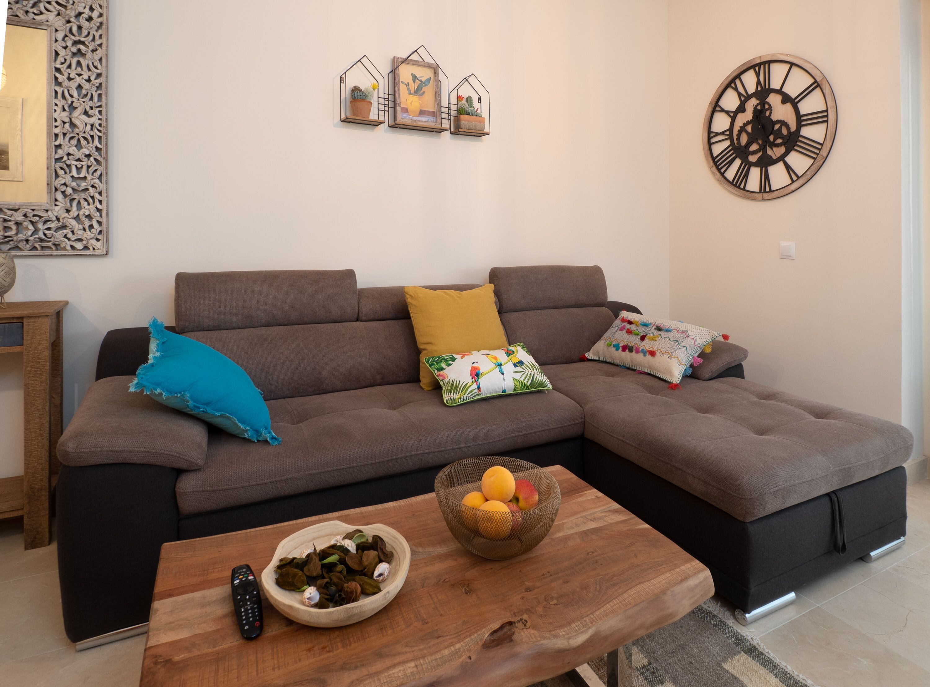 A modern living room with a sectional sofa, decorative pillows, a wooden coffee table with fruit and nuts, a wall clock, and framed cactus artwork.