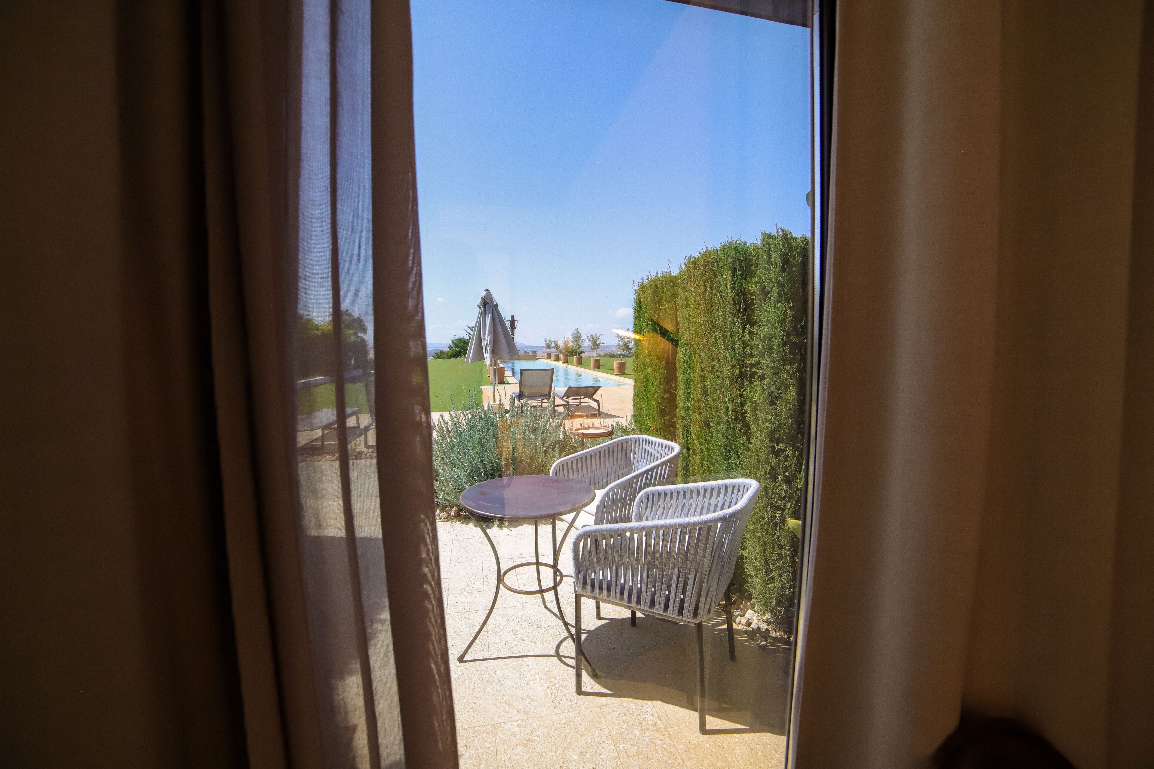 Blick durch ein Fenster auf einen Patio mit Pool, zwei weißen Stühlen und einem Tisch.