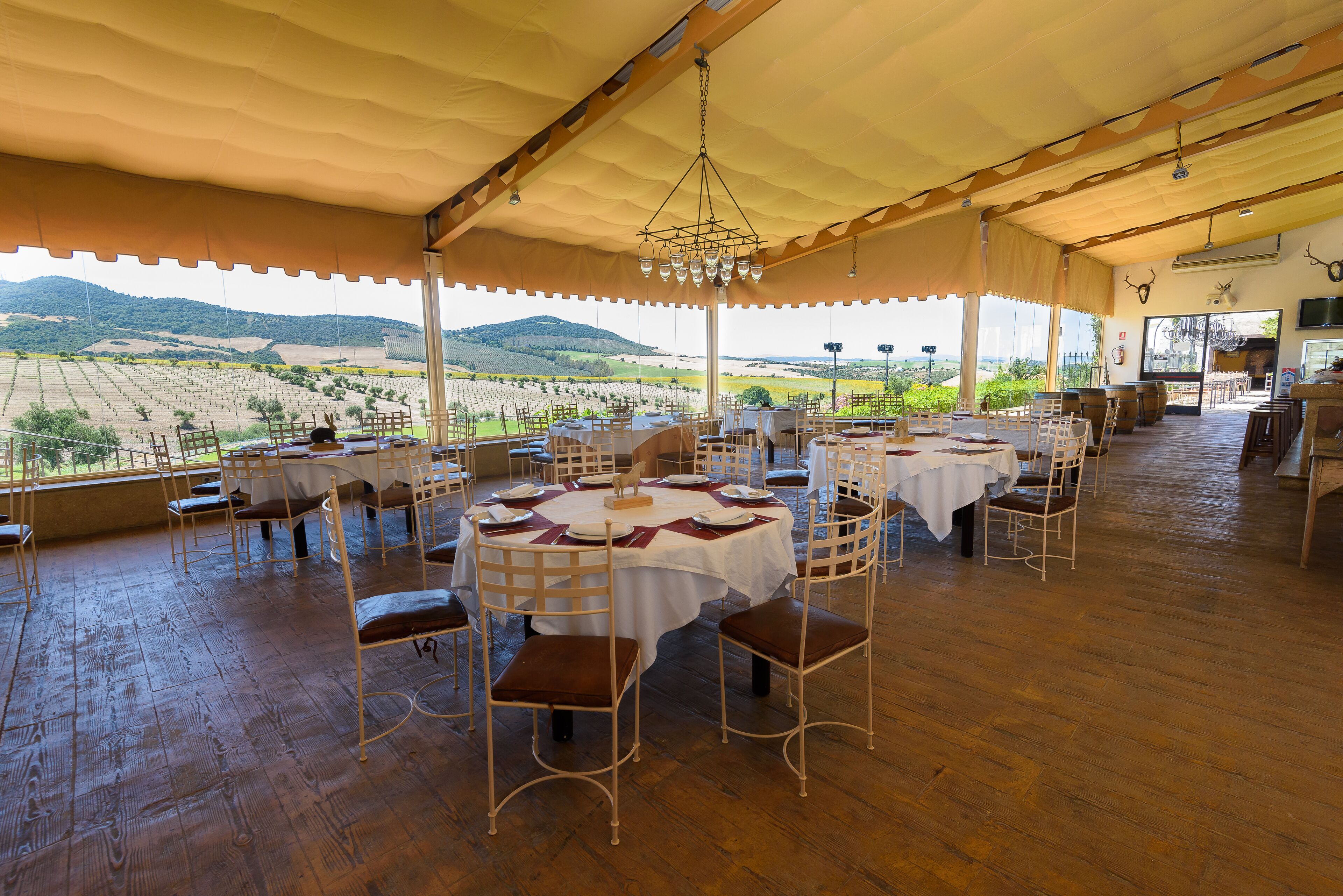An outdoor dining area with round tables and chairs, a wooden floor, and a view of the countryside.