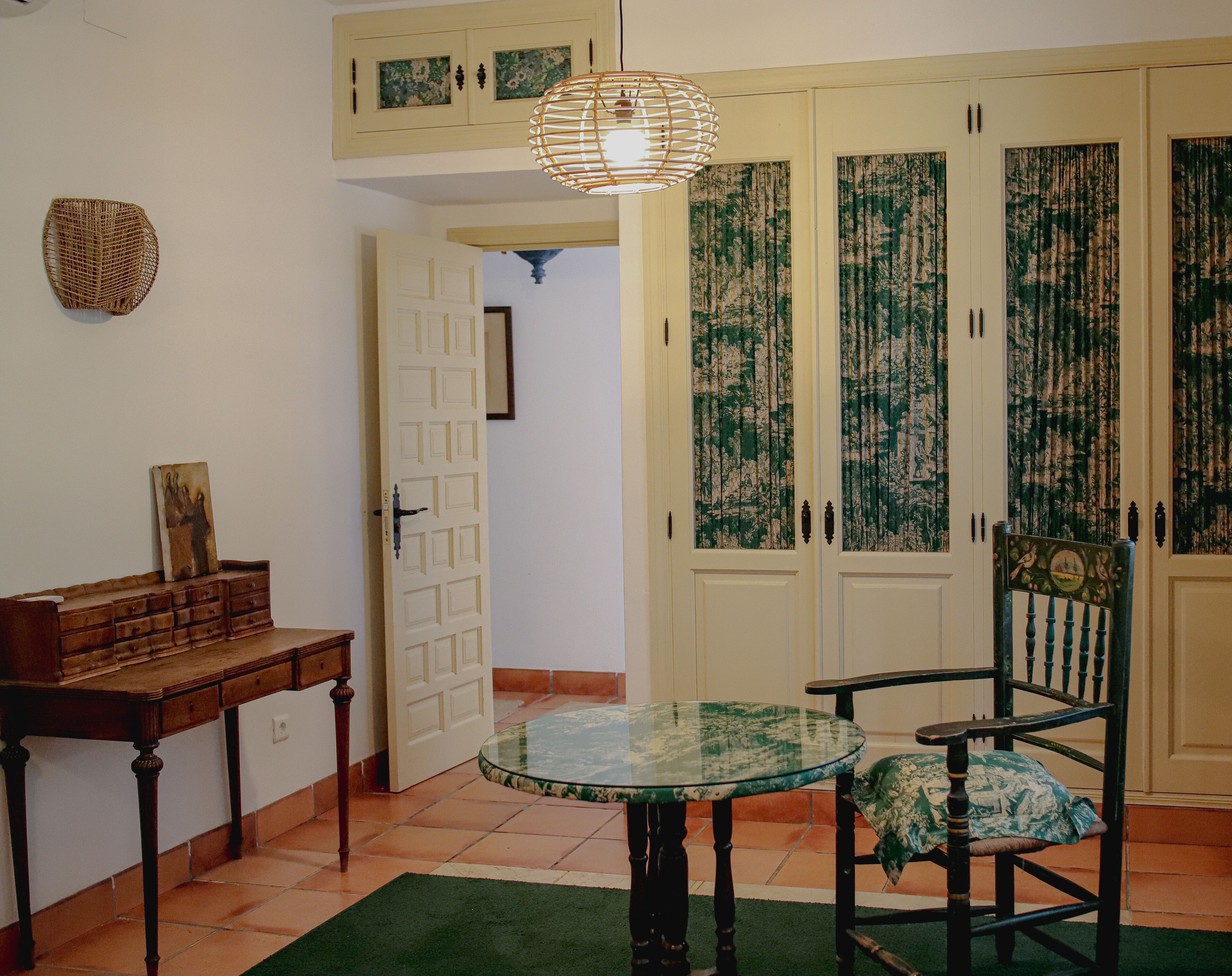 Una habitació amb una taula rodona de vidre, una cadira de fusta, un armari i una porta amb cortines florals.