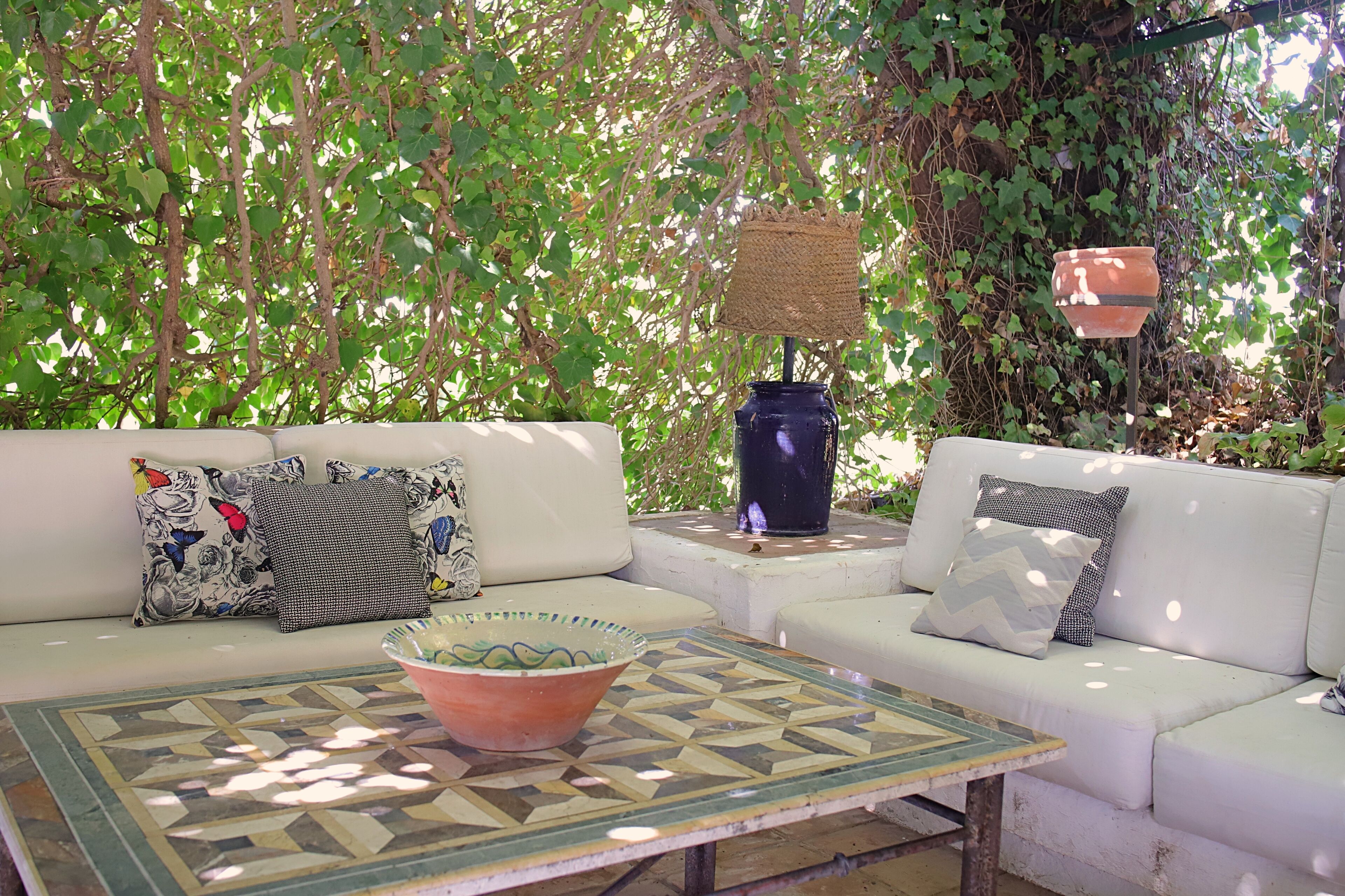 Un área de estar al aire libre con dos sofás blancos, cojines estampados, una mesa baja de baldosas y una maceta colgante.