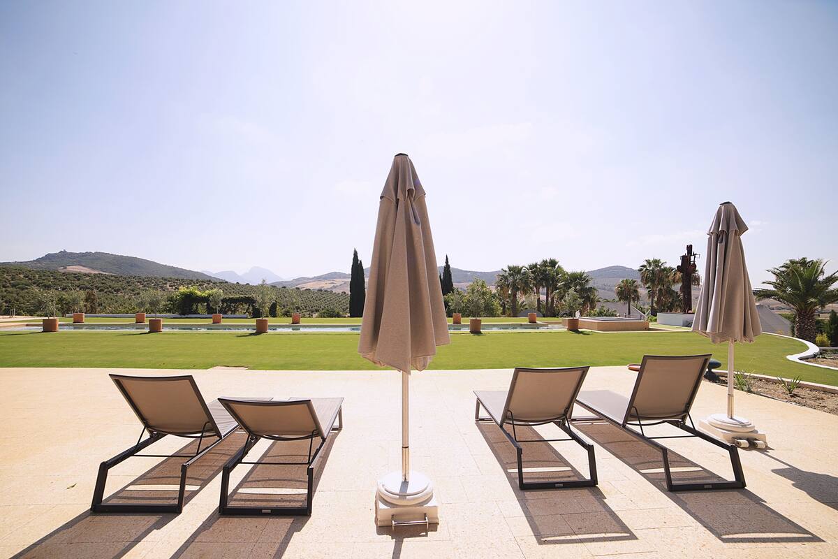 Un espace aménagé au bord de la piscine, avec des chaises longues et des parasols, une pelouse bien entretenue et des montagnes qui se dessinent à l’horizon.