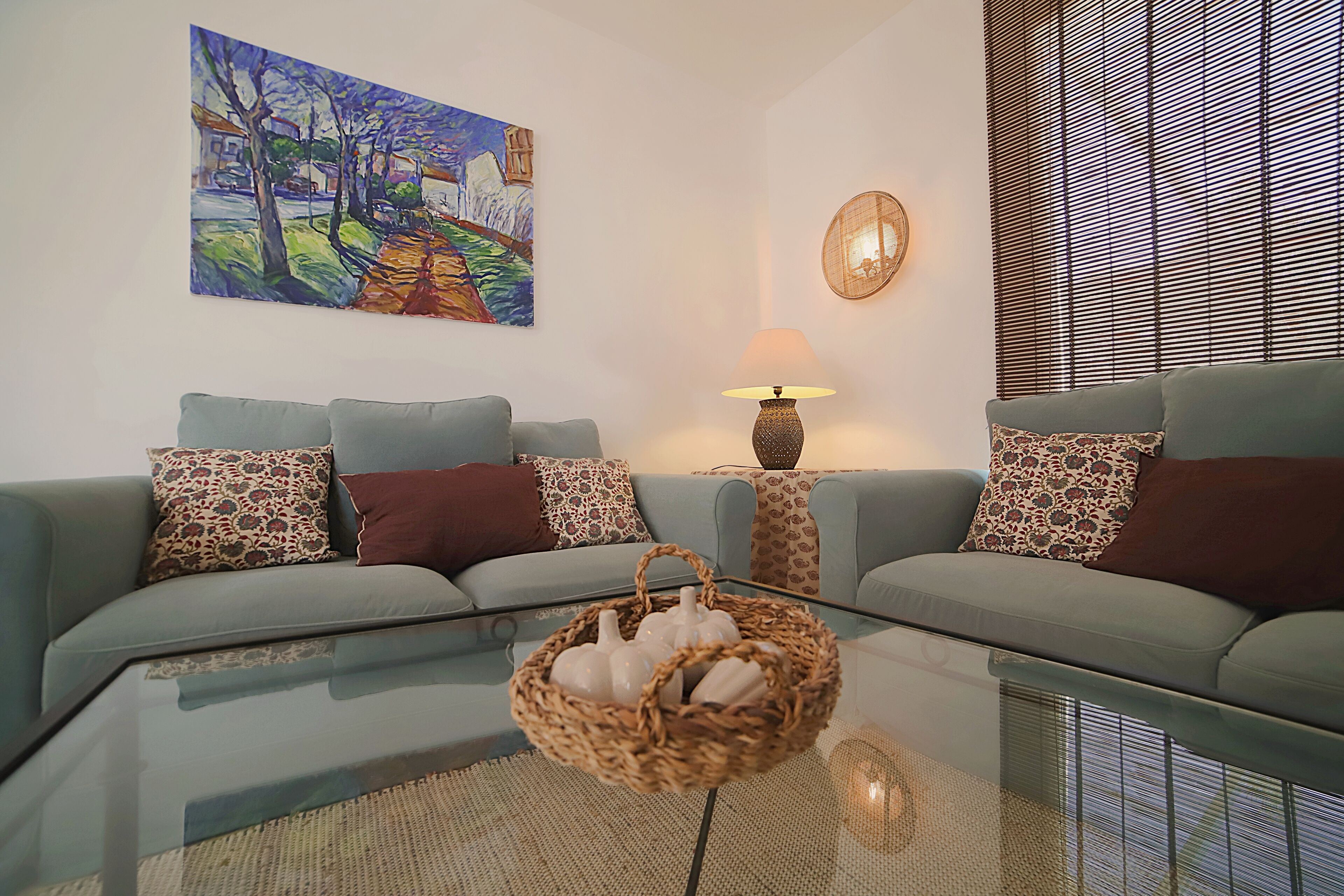 A living room with a sofa set, a glass coffee table, a painting on the wall, a lamp, and a window with blinds.