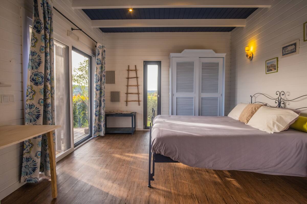 A bedroom with a bed, a wooden floor, a window with curtains, a door, and a ladder.