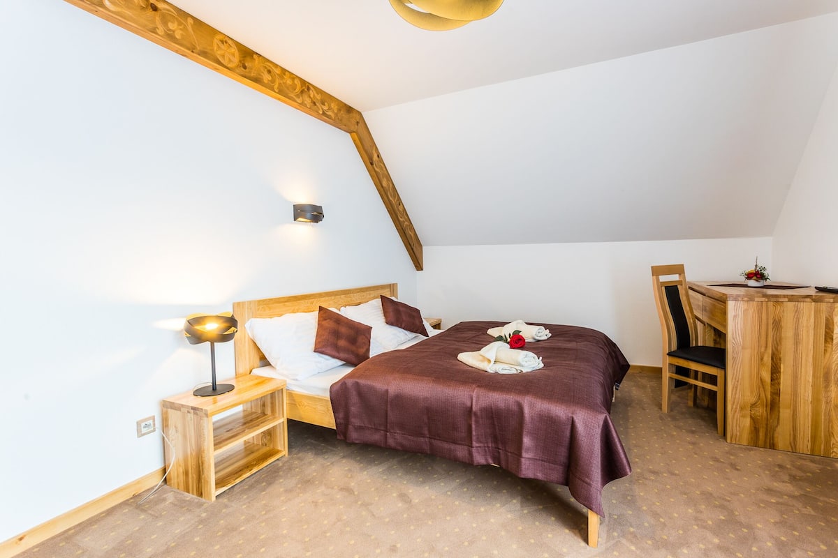Une chambre à coucher avec une tête de lit en bois, un lit recouvert d’une couvre-lit marron foncé, une lampe de chevet, une table de chevet en bois, une petite table avec une chaise et un luminaire fixé au mur.