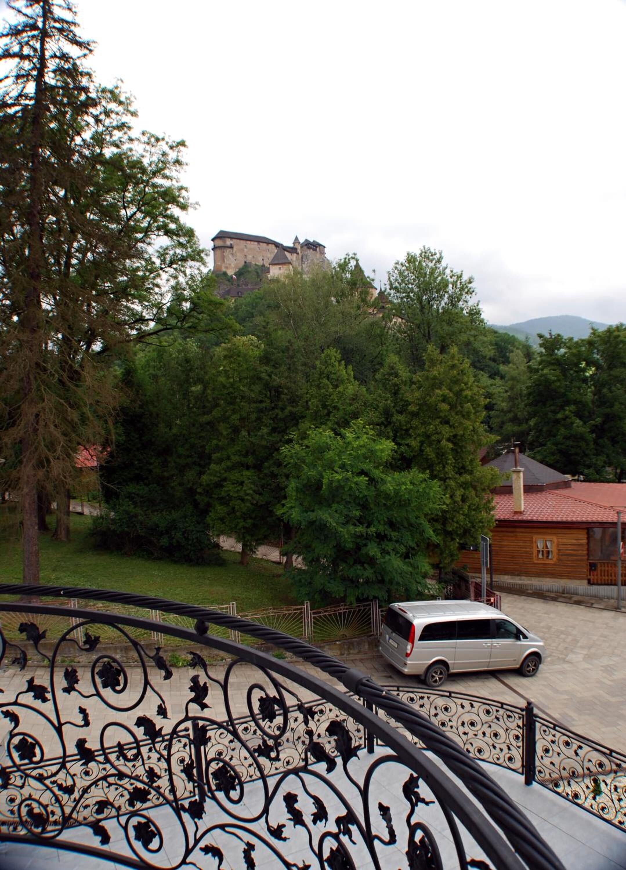 A view of a castle on a hill, surrounded by trees and a white van parked nearby.
