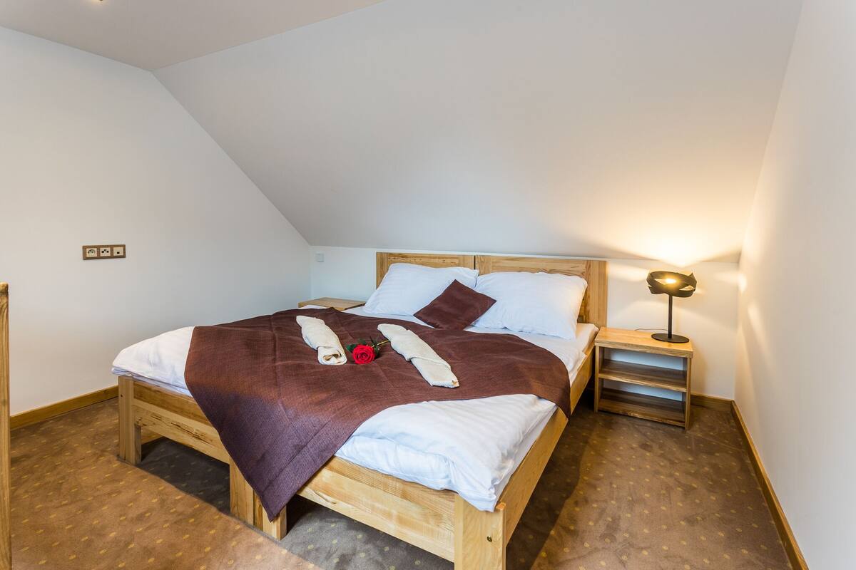 Un lit bien fait, recouvert d’une couverture marron, avec des oreillers blancs et une rose rouge. On y trouve une petite table de chevet avec une lampe et une commode en bois.