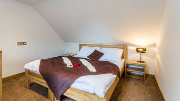 A neatly made bed with a brown blanket, white pillows, and a red rose. There is a small bedside table with a lamp and a wooden nightstand.