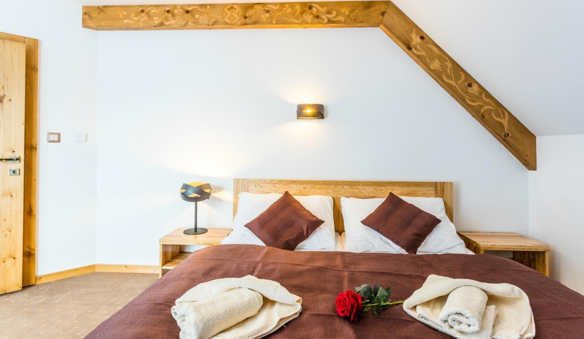 Een slaapkamer met een schuine kap, een houten hoofdbord, twee nachtkastjes, een lamp, een bed met twee kussens en handdoeken op het bed.
