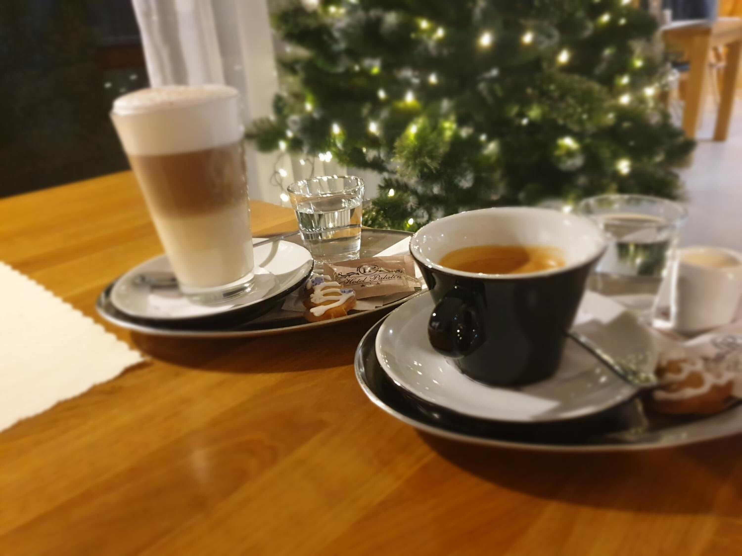 A table with a coffee cup, a glass of water, a plate with a pastry, and a small Christmas tree in the background.
