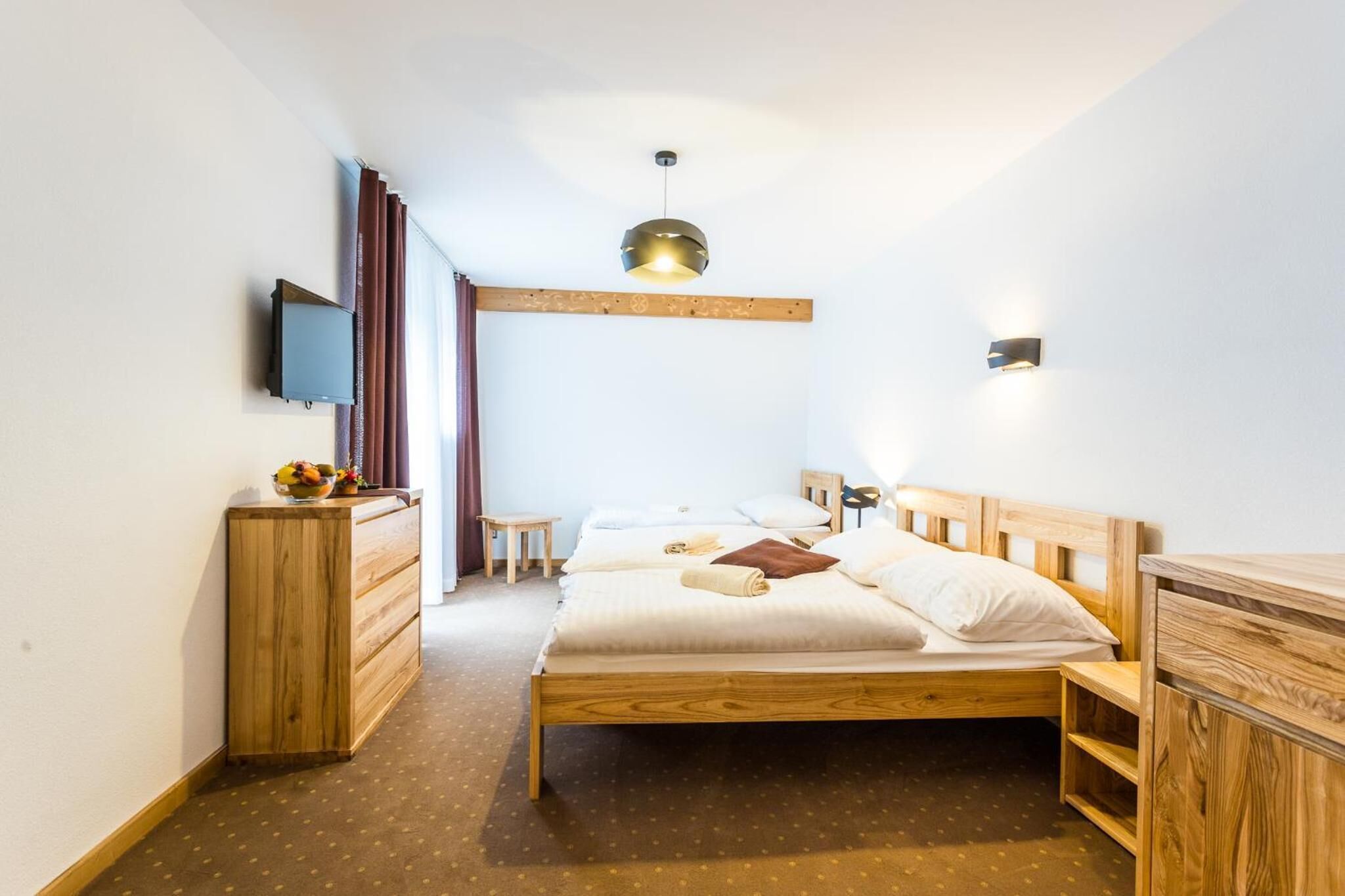 A bedroom with a wooden bed, a small wooden dresser, a television mounted on the wall, and a pendant light.