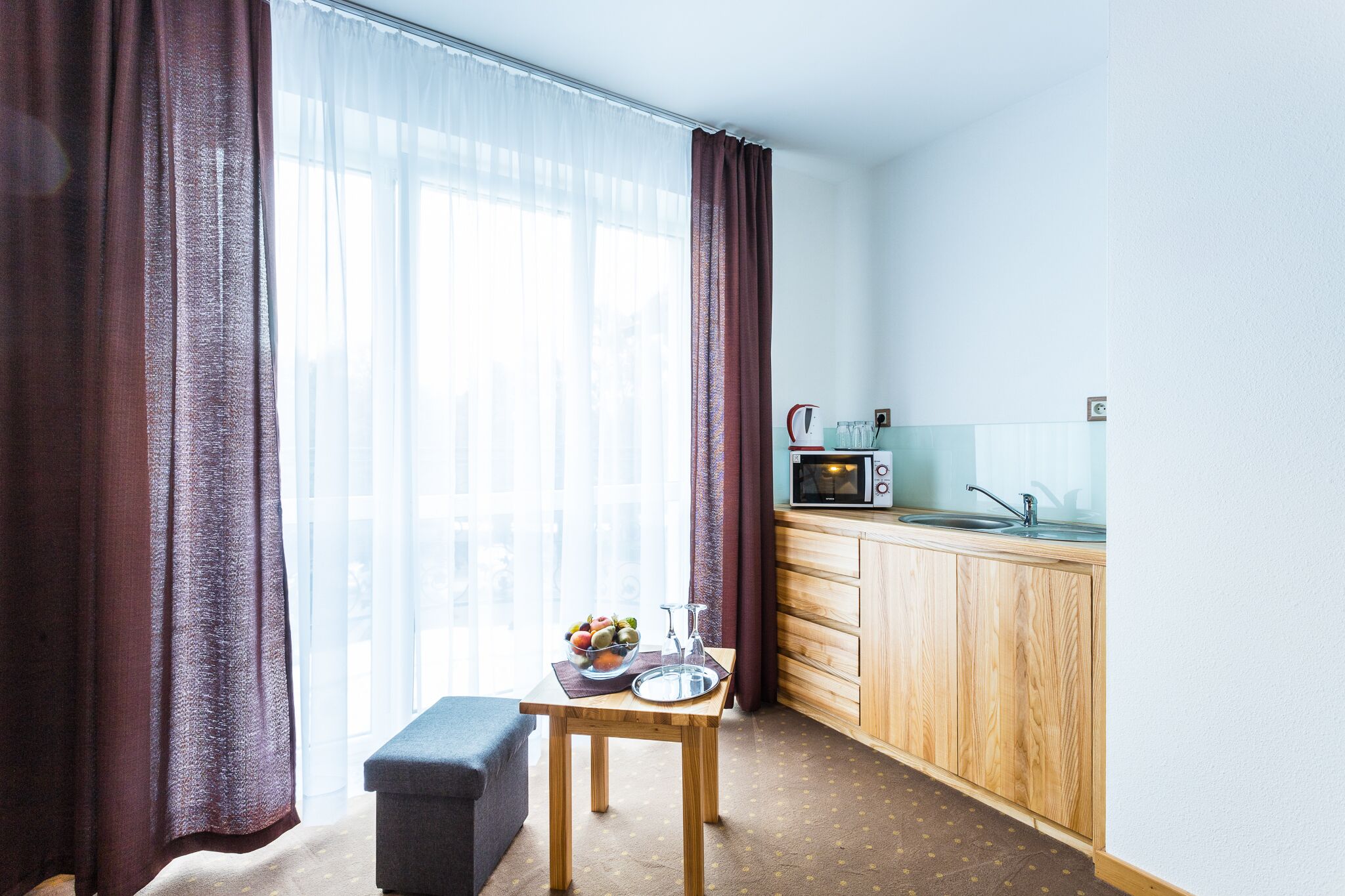 A small kitchenette with a microwave, sink, and wooden cabinets. A table with a fruit bowl and a glass is in front of a window with sheer curtains.