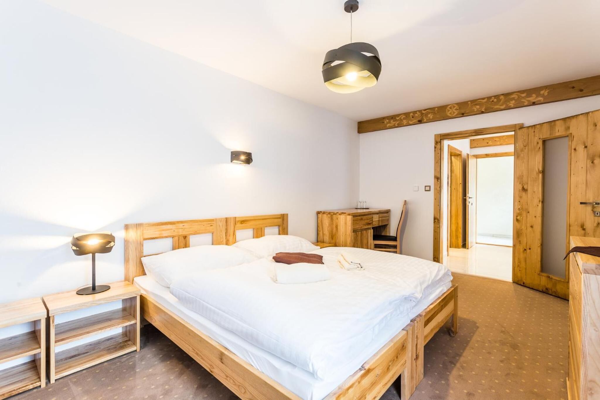 Ein hölzernes Bett mit weißer Matratze, ein Nachttisch mit Lampe sowie ein Holztisch mit Stuhl.