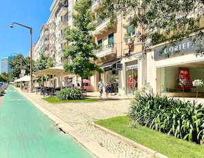 Una calle con un carril bici verde, mesas al aire libre y locales comerciales, incluyendo un cartel visible que dice 'CAROL'.