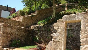 Um edifício de pedra com uma porta, rodeado por vegetação e um muro de pedra.