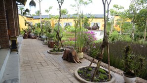 Un sendero pavimentado bordeado de macetas con plantas y árboles, que conduce a un edificio amarillo con techo de tejas.