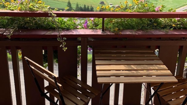 En träveranda med två stolar och ett bord, med utsikt över en dal med gröna fält och berg i bakgrunden.
