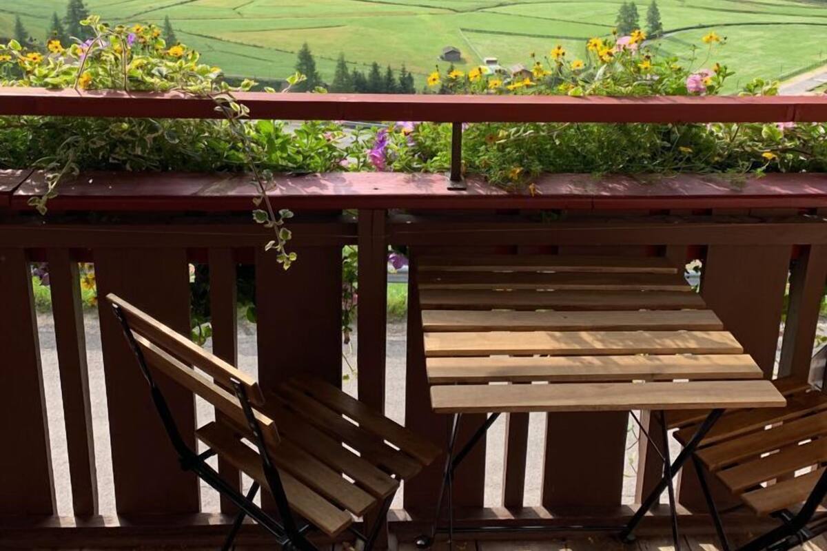 Une terrasse en bois avec deux chaises et une table, d’où l’on peut admirer une vallée verdoyante, avec des champs et des montagnes en arrière-plan.