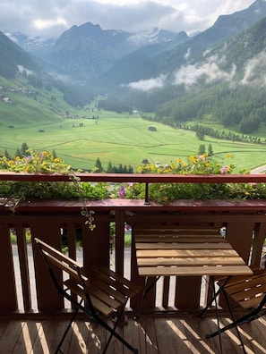 A wooden deck with two chairs and a table, overlooking a valley with green fields and mountains in the background.