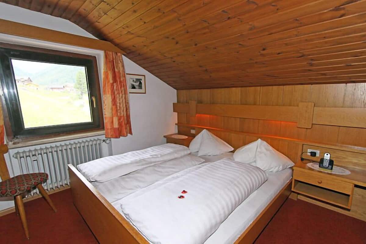 Une chambre avec un plafond en bois, une grande fenêtre avec des rideaux, un lit avec du linge de lit blanc, une table de chevet avec un téléphone et une chaise avec un coussin à motifs.