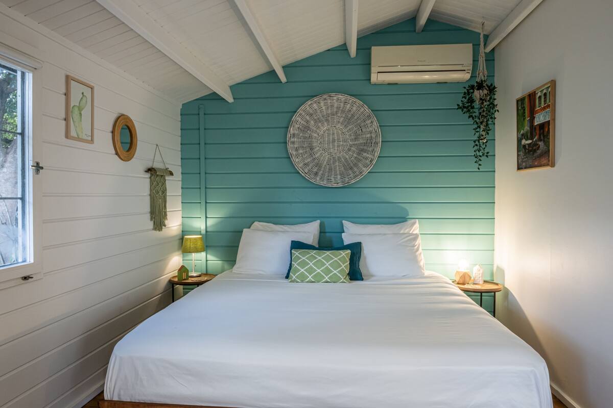 A bedroom with a bed, a window, a wall-mounted air conditioner, and a hanging plant.