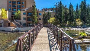Eine Holzbrücke führt zu einem mehrstöckigen Hotel mit Pool und Blick auf eine bewaldete Landschaft.