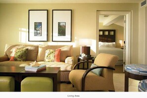 A living area with a sofa, armchairs, a coffee table, and a side table with a lamp. There are framed pictures on the wall and a bed in the background.