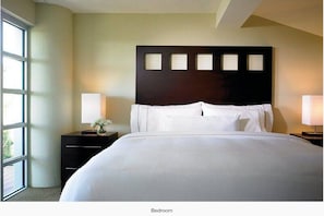 Une chambre avec un grand lit, une tête de lit sombre ornée de quatre découpes carrées, deux tables de chevet avec des lampes et une fenêtre sur le côté gauche.