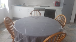 Et køkken med hvide skabe, en vask, opvaskemaskine, mikroovn og køleskab. Der er et rundt bord med stole og en plastikdug.