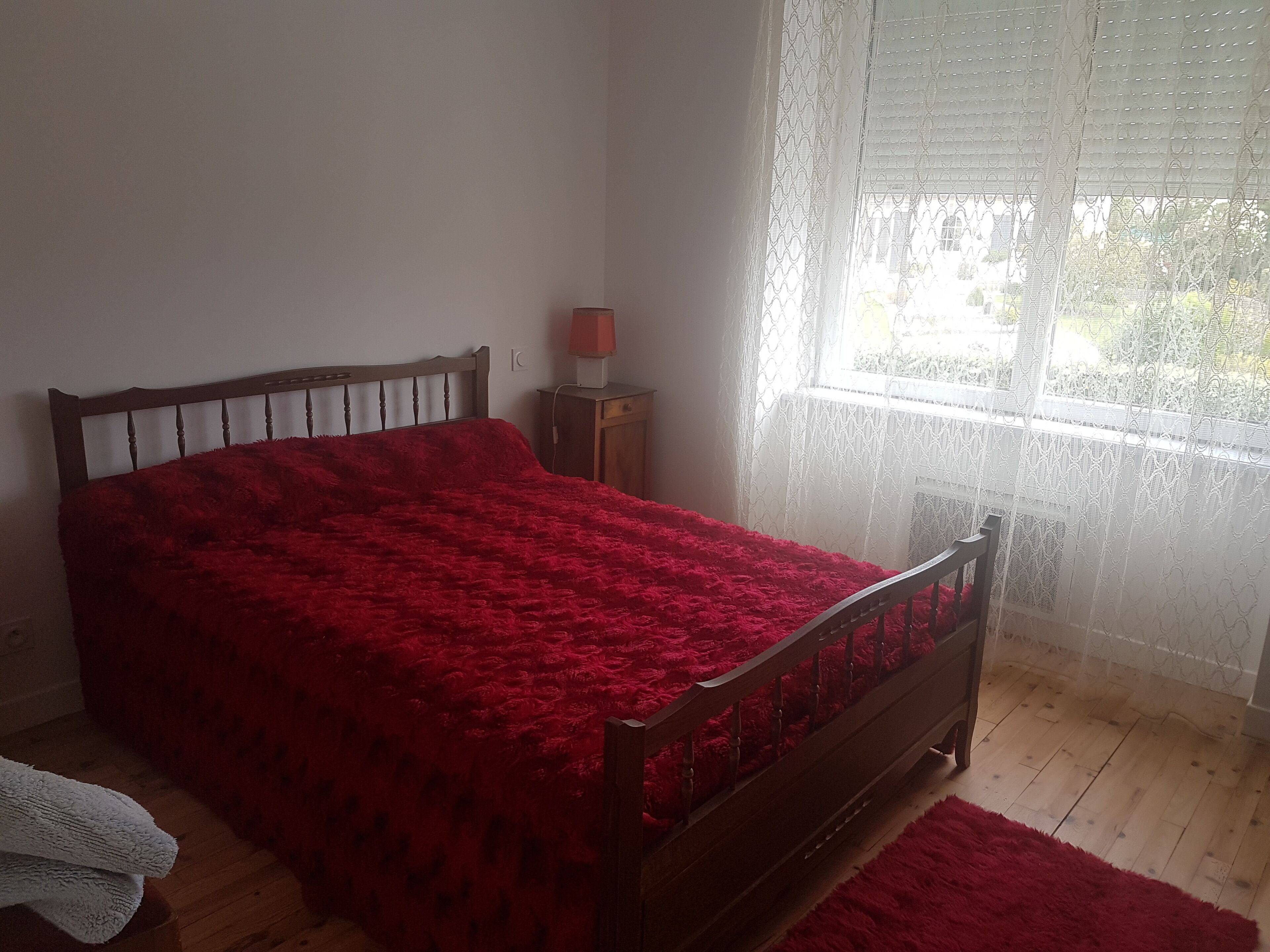 A bedroom with a wooden bed, a red blanket, a nightstand with a lamp, and a window with sheer curtains.