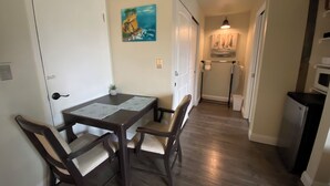 Un coin repas avec une table en bois et quatre chaises, un tableau accroché au mur, et un couloir menant à la cuisine.