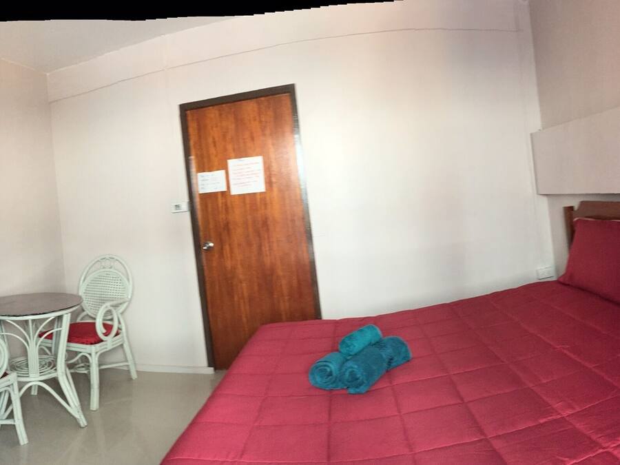 Una habitación pequeña y compacta con una cama individual, una puerta de madera, una mesita con dos sillas y una colcha roja.