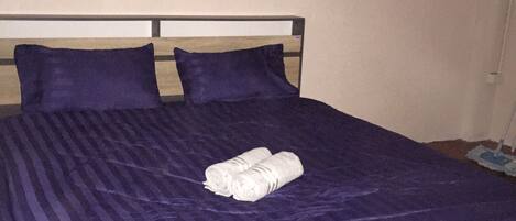 A bed with a purple bedspread and two pillows, a folded towel on the bed, and a mop leaning against the wall.