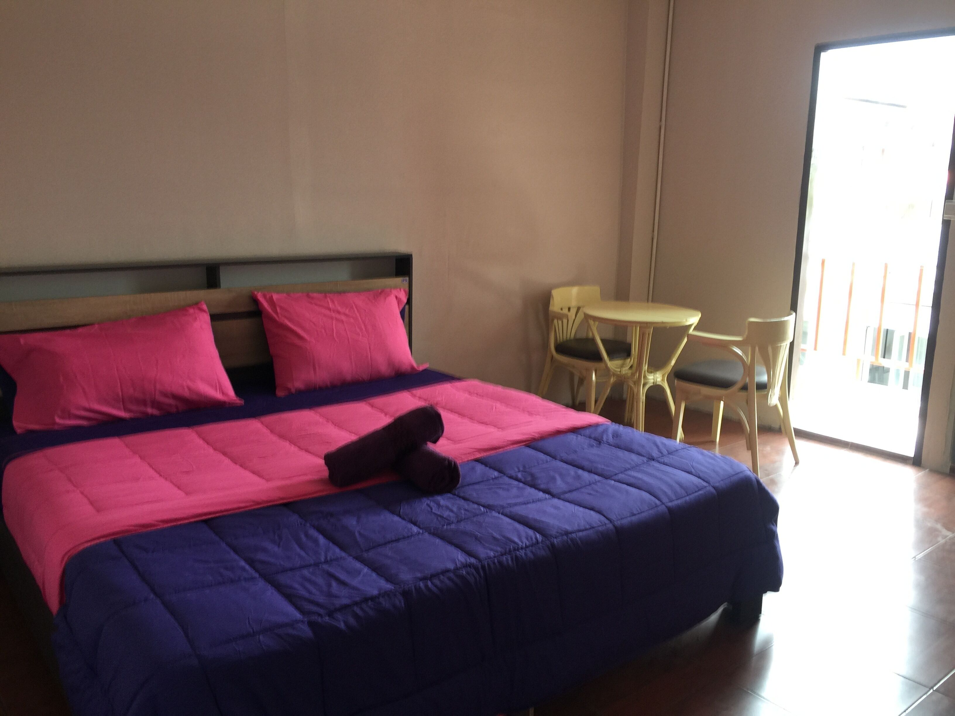 Una cama con una colcha azul y rosa, dos almohadas y un par de zapatillas. En la esquina hay una mesita y dos sillas.