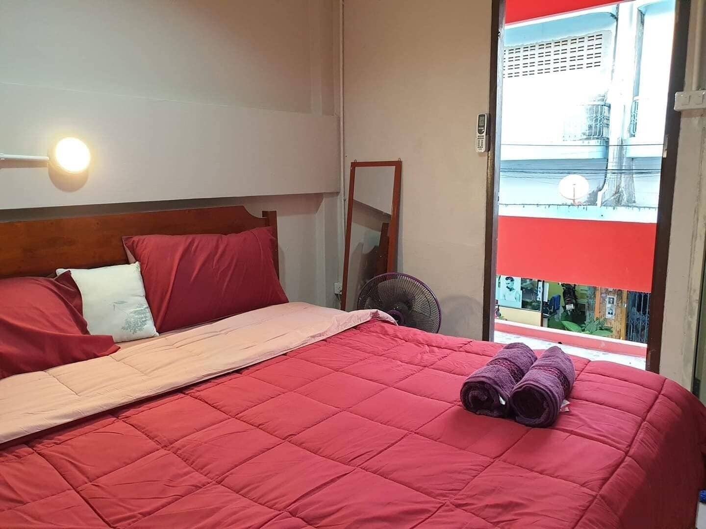 A bed with red bedding, a bedside fan, and a window with a view of buildings.