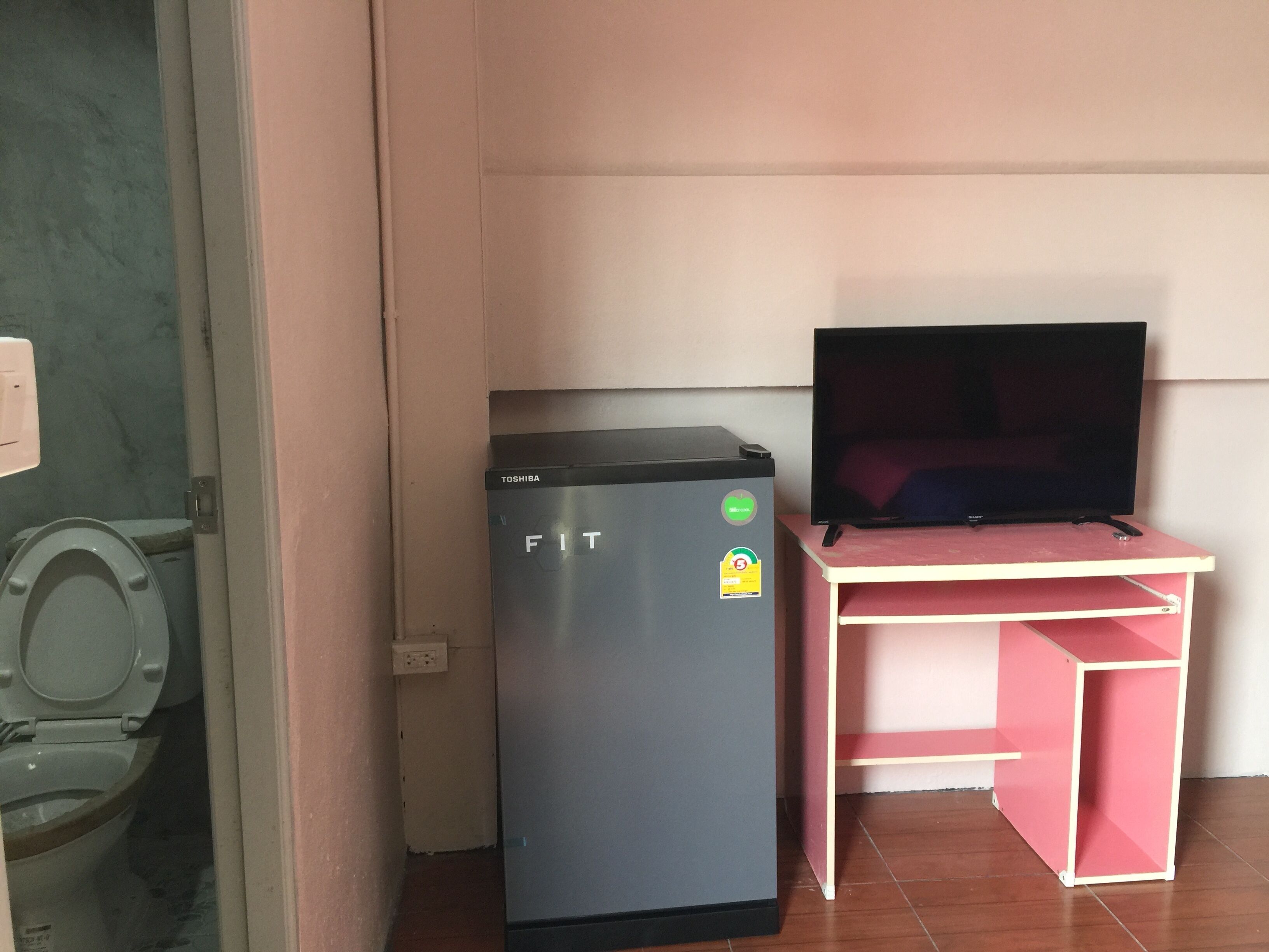 A compact room with a refrigerator, a television on a pink desk, and a toilet.