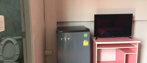 A compact room with a refrigerator, a television on a pink desk, and a toilet.