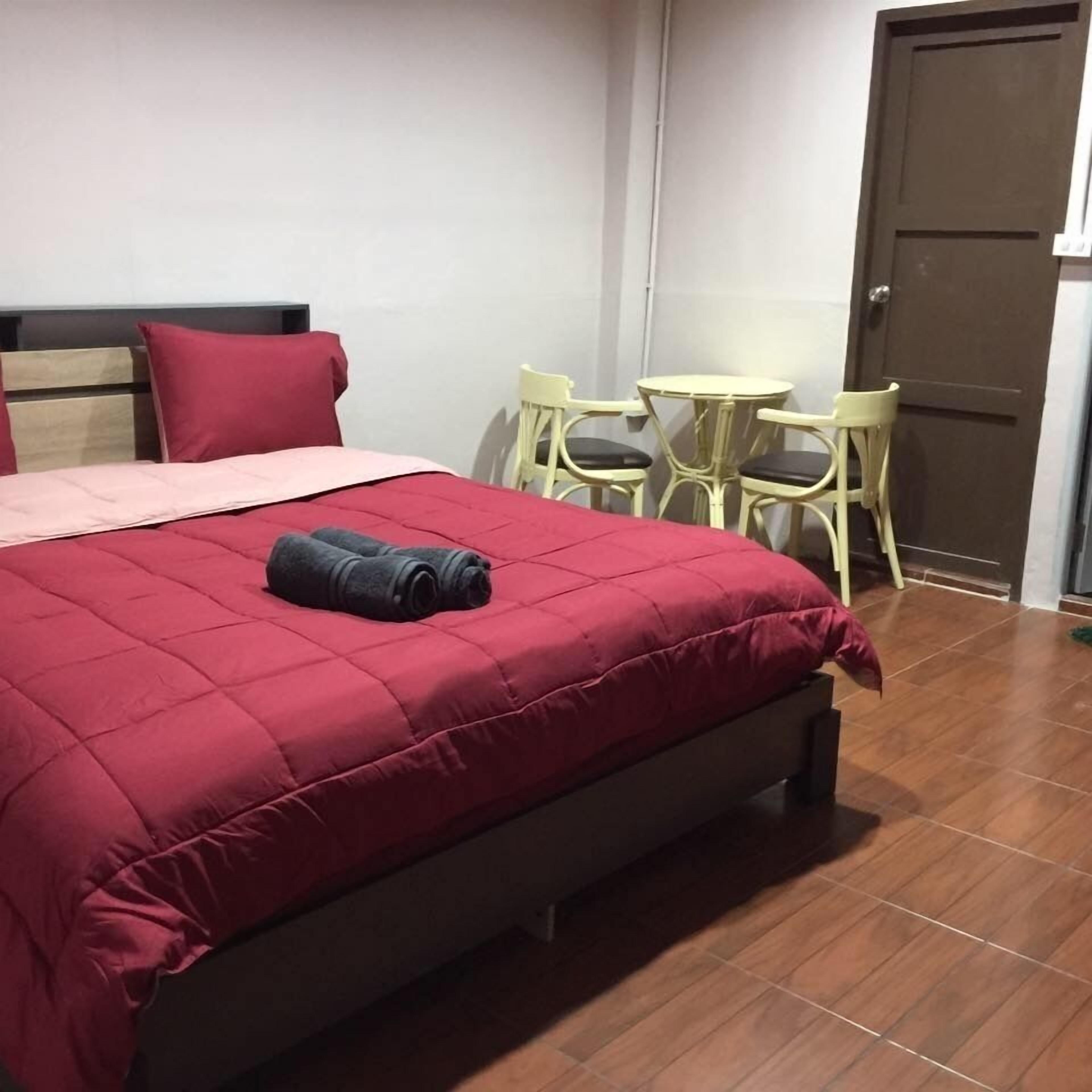 Una cama bien hecha con colcha roja, una mesita redonda con sillas y una puerta al fondo.