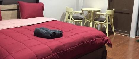 Una cama bien hecha con colcha roja, una mesita redonda con sillas y una puerta al fondo.