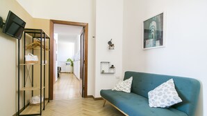 Un salon moderne avec un canapé de couleur turquoise, un plancher en bois, un téléviseur fixé au mur et un couloir menant à la cuisine.
