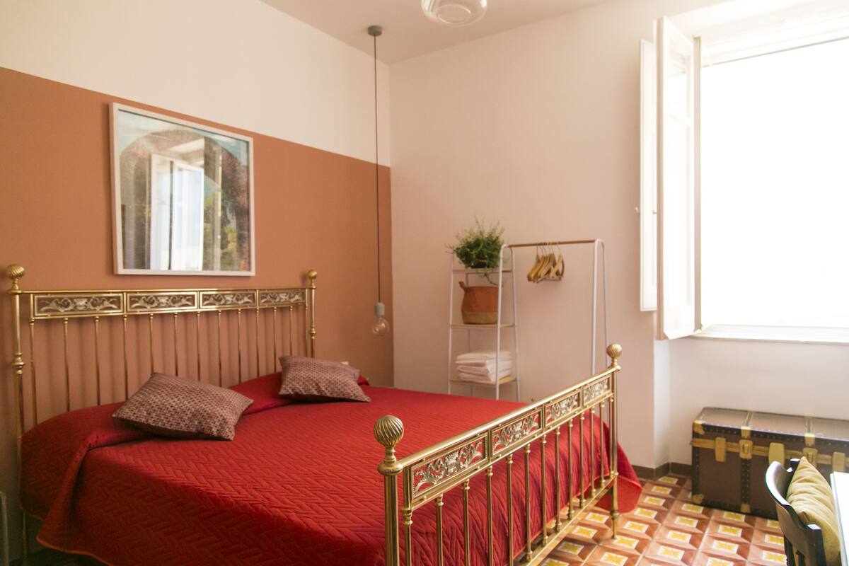 Une chambre à coucher avec un lit, une fenêtre, une plante et une valise.