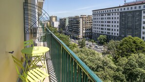Un balcon doté d’une balustrade verte, d’une table jaune et de chaises, offrant une vue sur un paysage urbain avec des bâtiments et des arbres.
