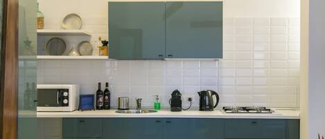 A kitchen with white tiles, a microwave, a kettle, a coffee maker, and cabinets.