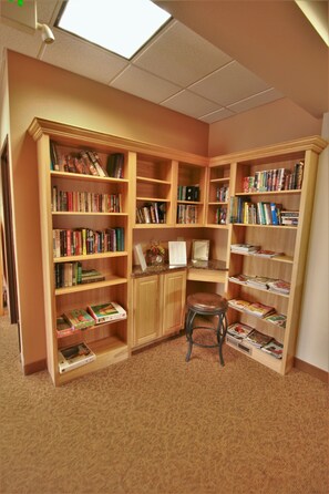 A library with multiple wooden bookshelves filled with books and magazines. There is a small wooden stool in the center.
