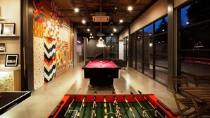 A billiards table in a room with a colorful wall mural, a foosball table, and large windows. - The Social House - Coliving & Hostel (Jakarta)