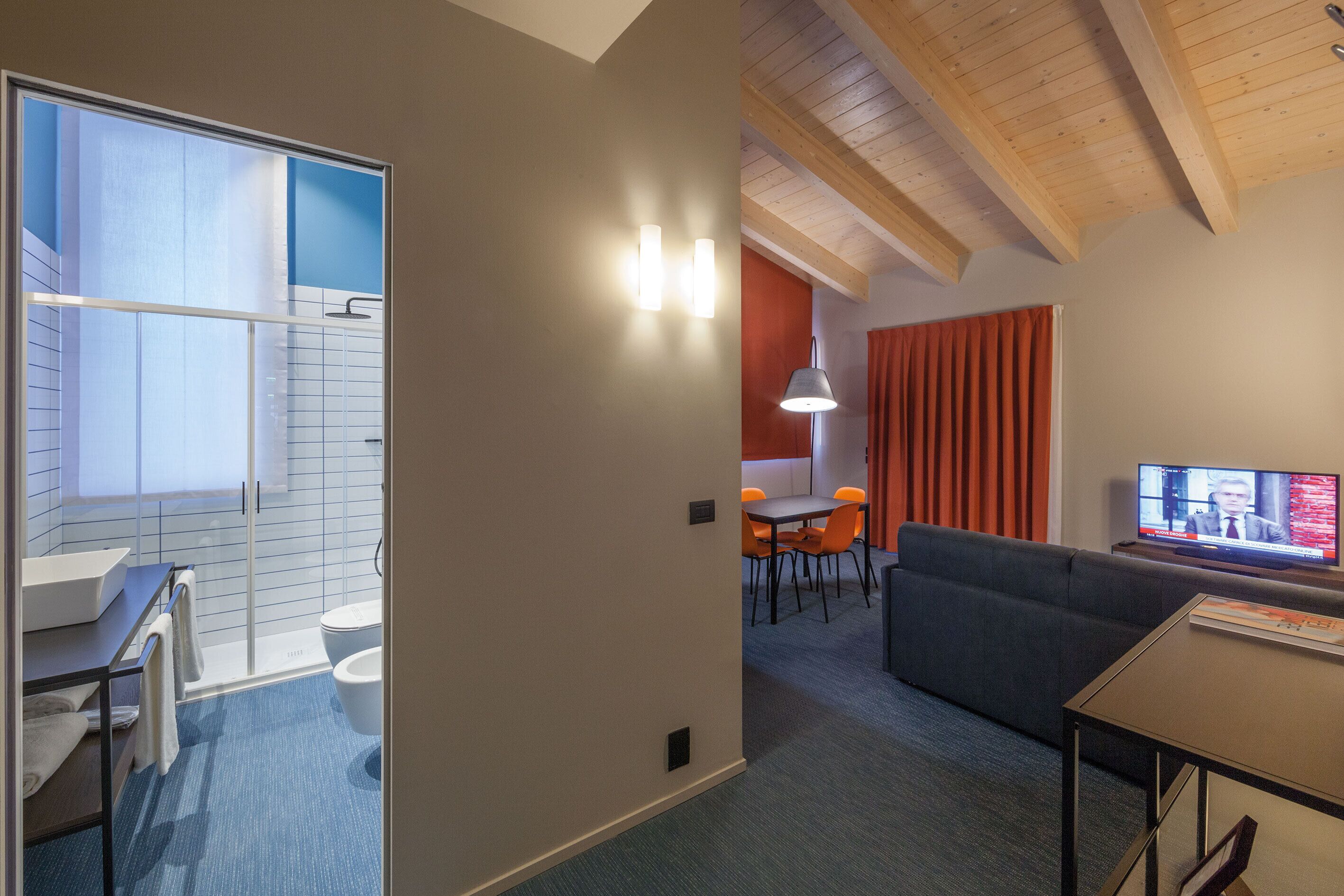 Habitación de hotel moderna con ducha, inodoro y zona de comedor con sillas naranjas.