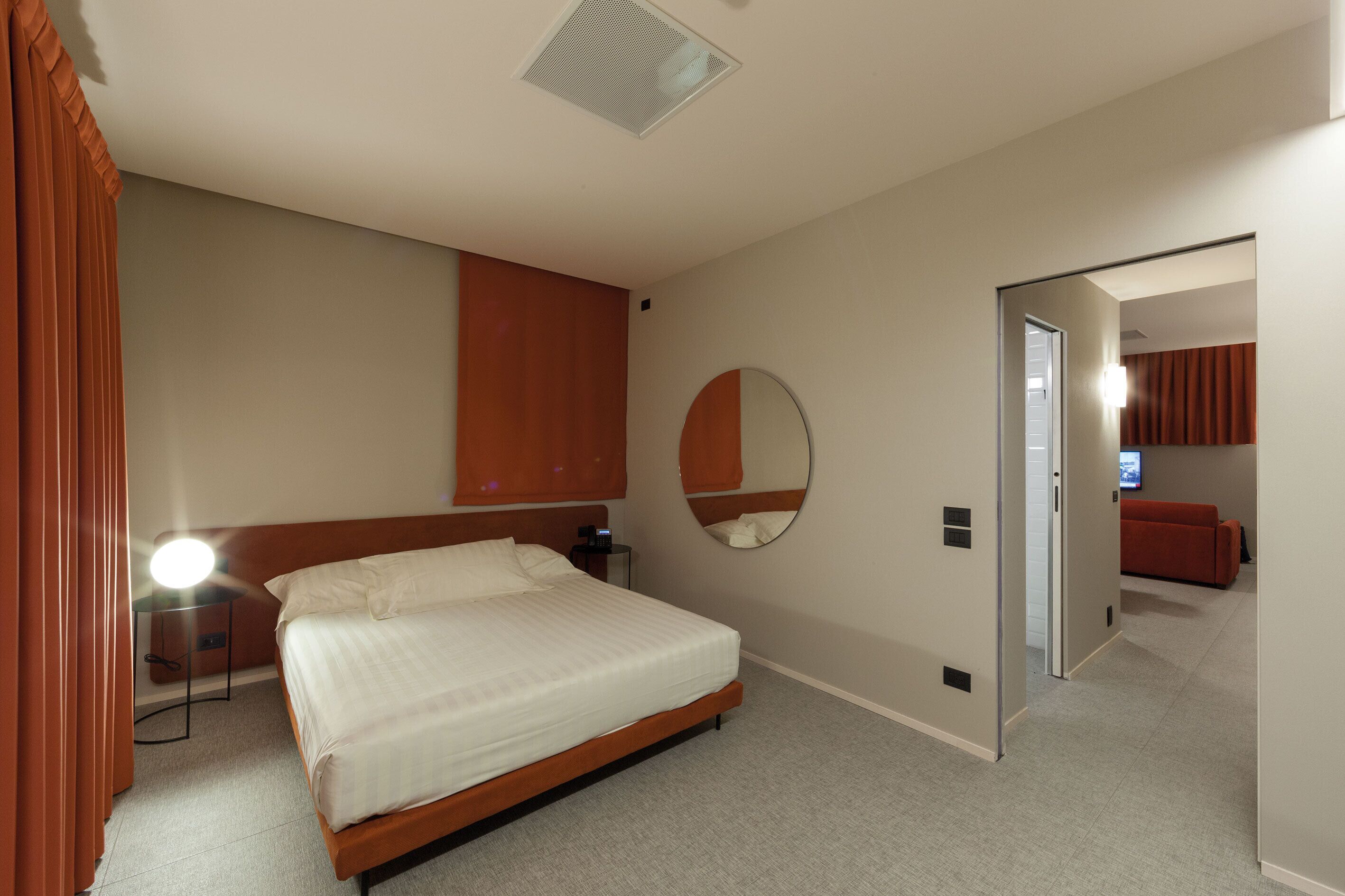 A hotel room with a bed, bedside table, lamp, mirror, and a door leading to another room.